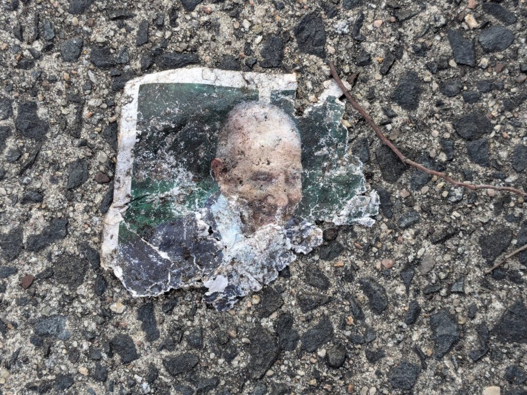 Color photograph looking down at a small, badly weathered printed portrait lying on coarse asphalt. The torn and dirt-stained photograph shows an older man in a dark jacket against a green background, though much of his face and lower body have been worn away or obscured by grime. Jagged stones and pebbles surround the photograph, with a thin brown twig curving alongside it.